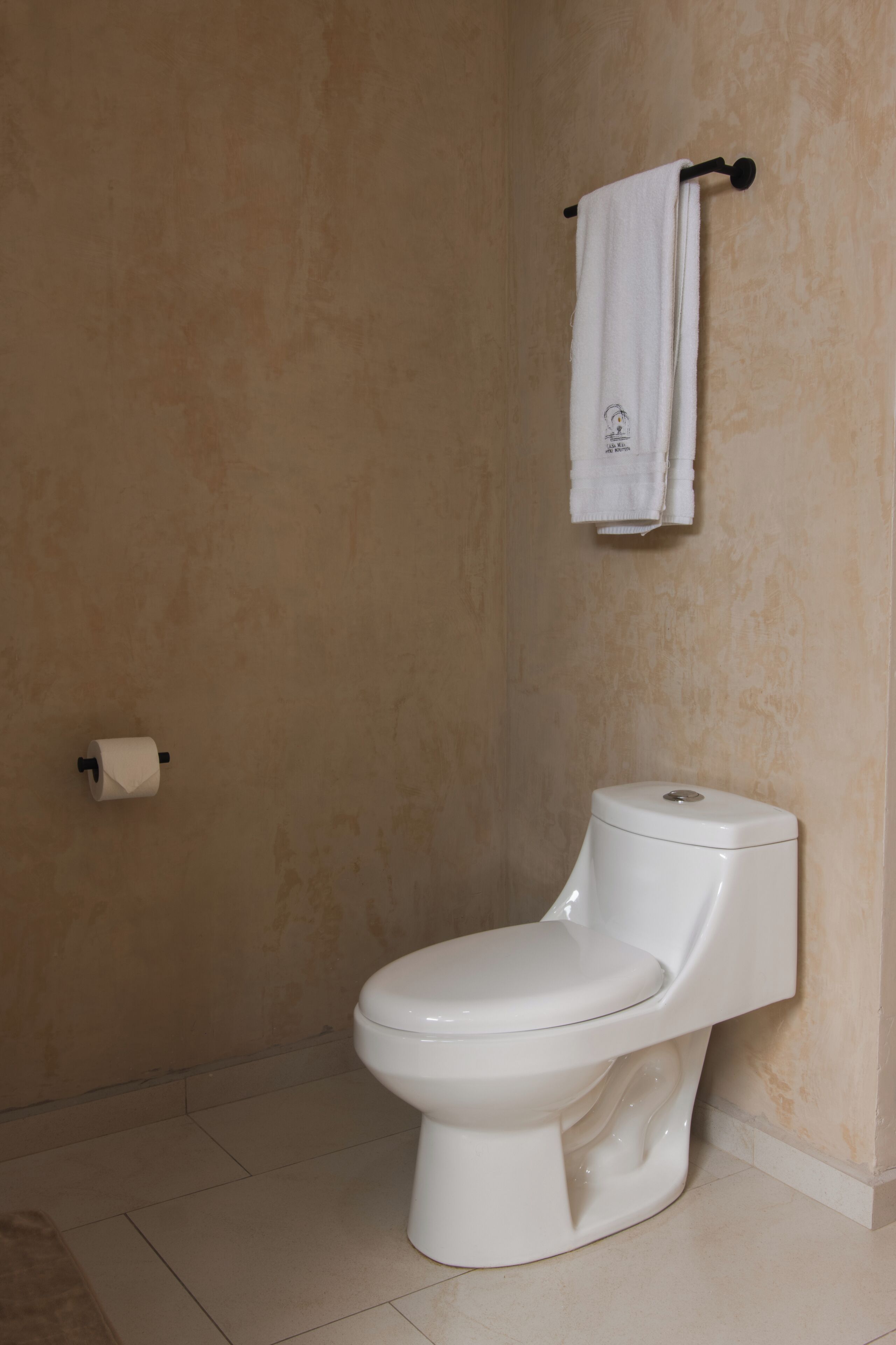A white toilet with a tank and seat, a roll of toilet paper on the floor, and a towel hanging on a hook on the wall.
