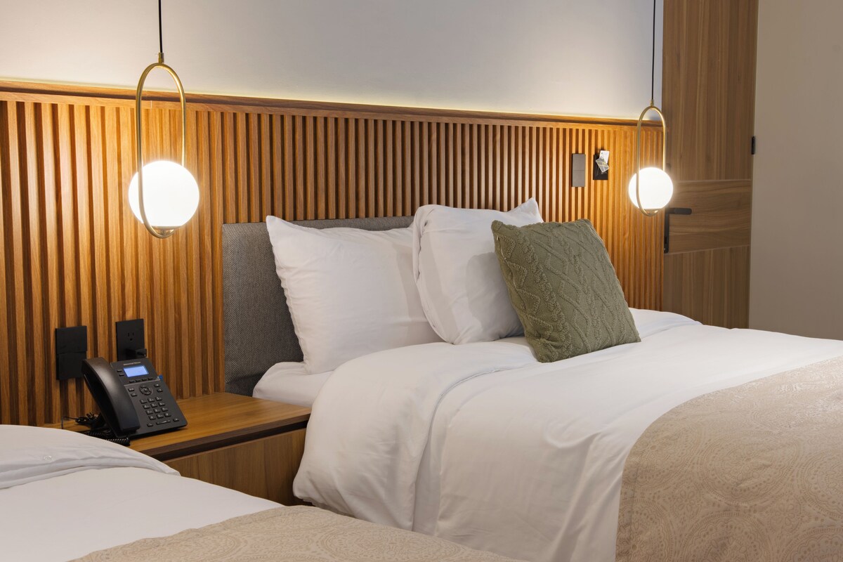 Une chambre d’hôtel avec une tête de lit en bois, deux lampes de chevet, un téléphone et un lit avec une literie blanche et plusieurs oreillers.