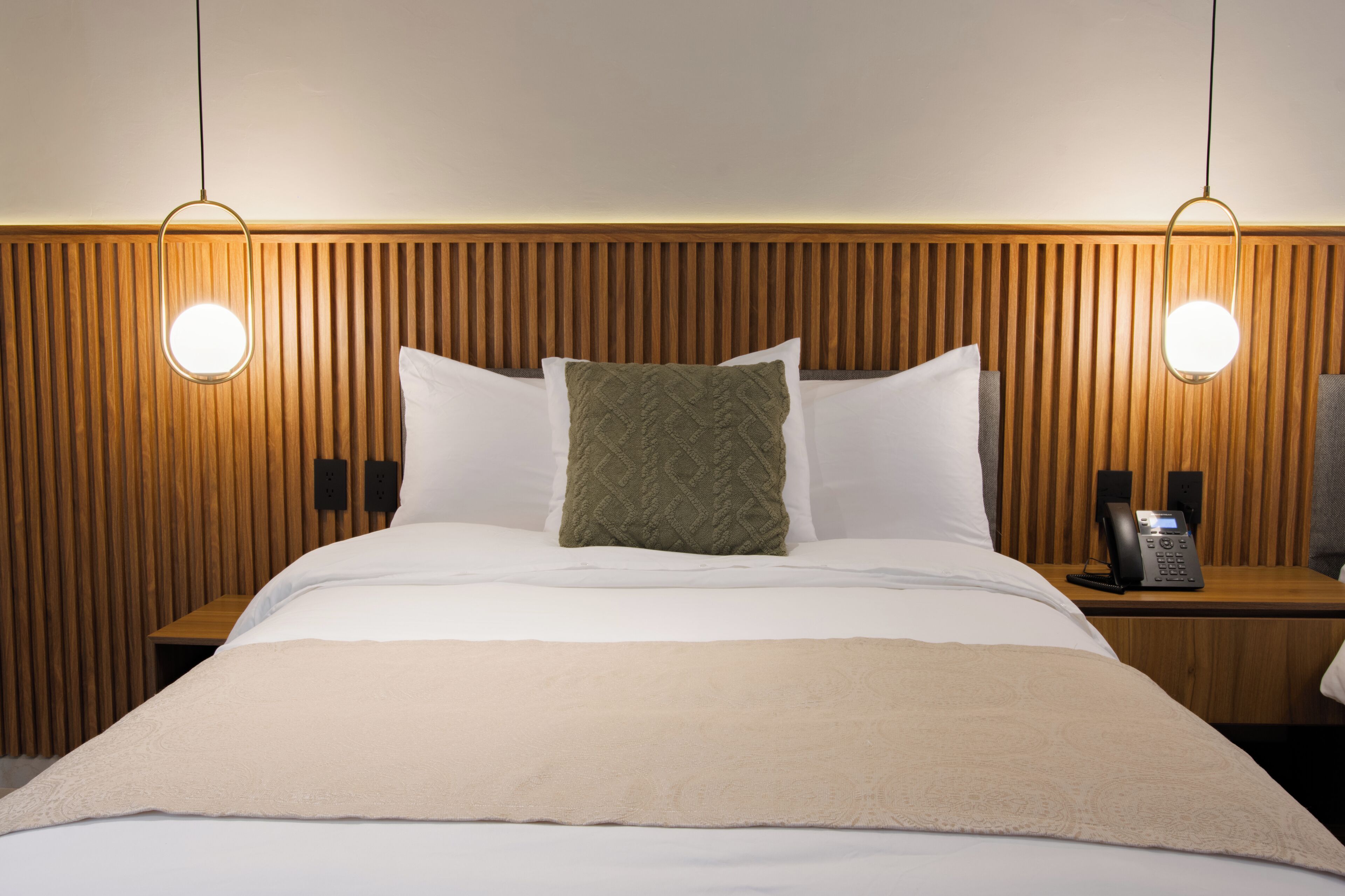 A hotel room with a wooden headboard, a bed with white linens, a green throw pillow, and a bedside table with a telephone.