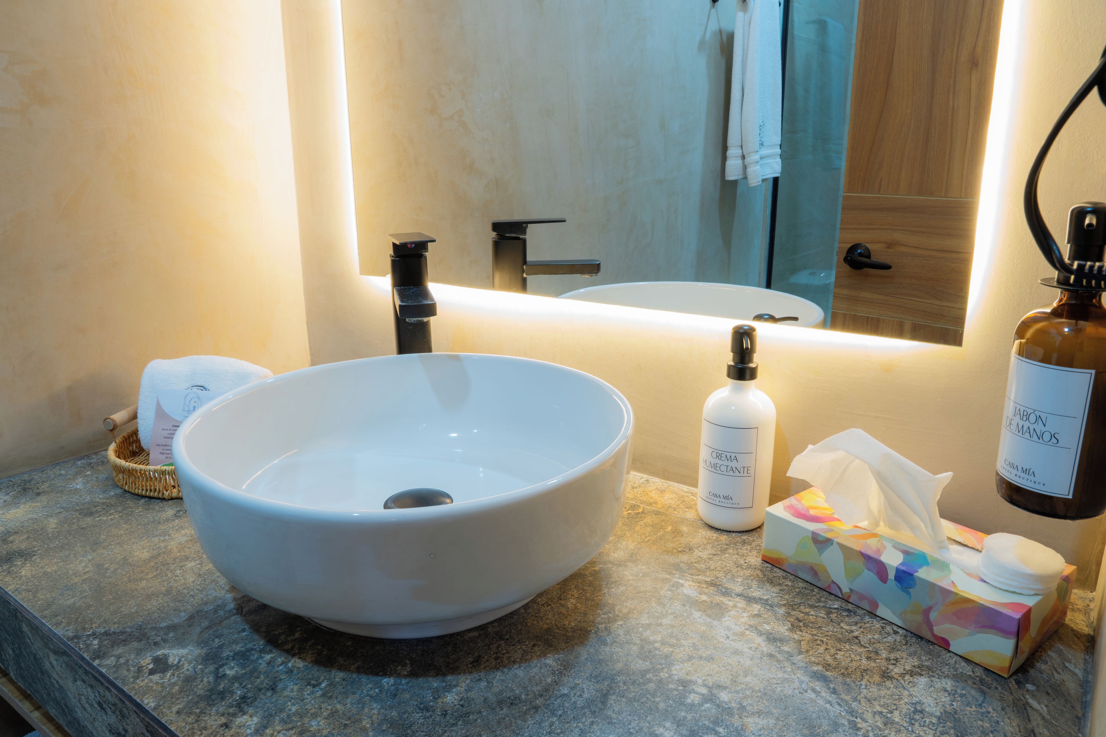 A bathroom with a large white sink, a mirror, a towel rack, a bottle of hand cream, and a tissue box.
