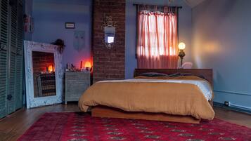 A bedroom with a brick fireplace, a bed with a red patterned rug, and a window with sheer curtains.