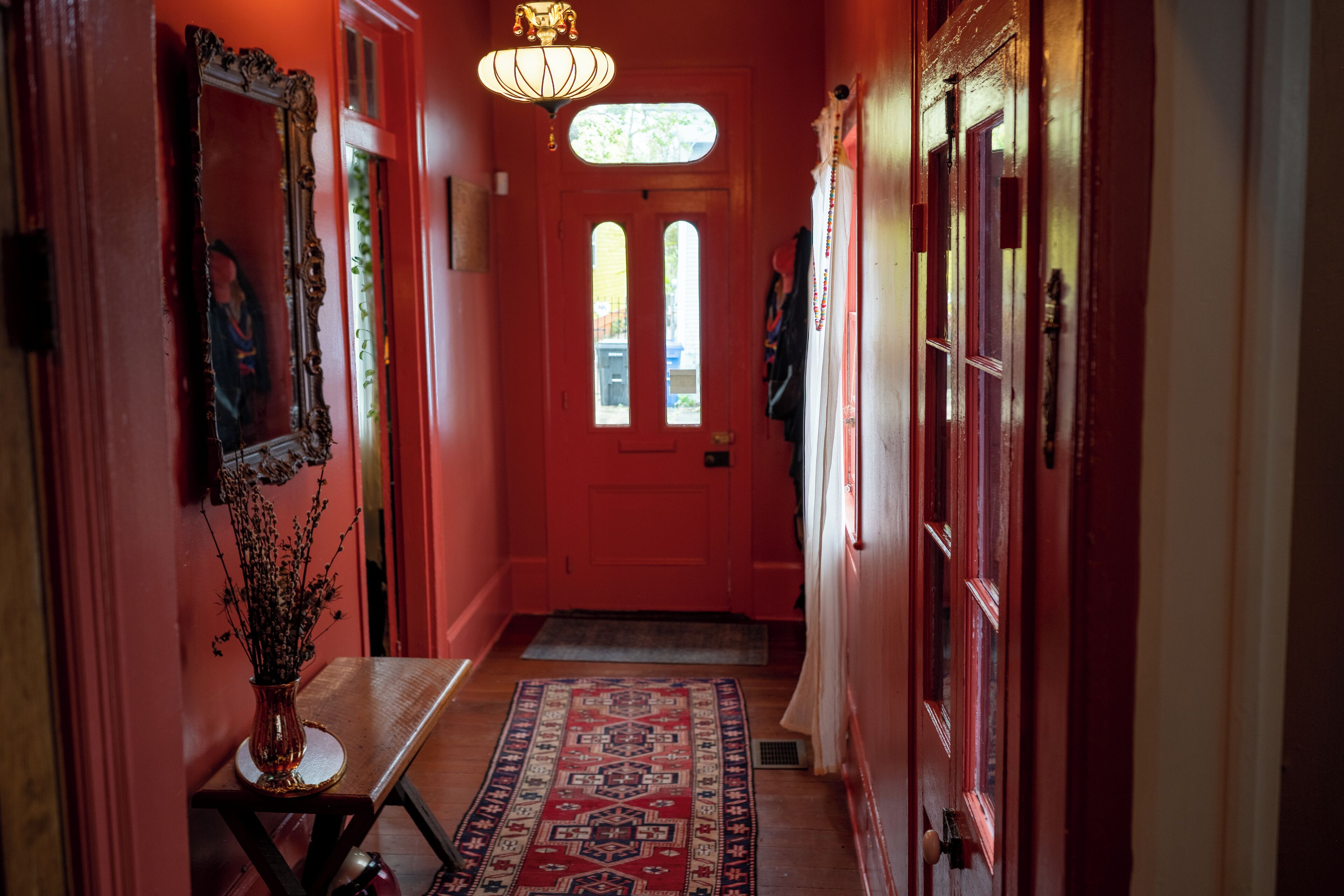 A hallway with a red door, a mirror, a vase with dried flowers, and a patterned rug.