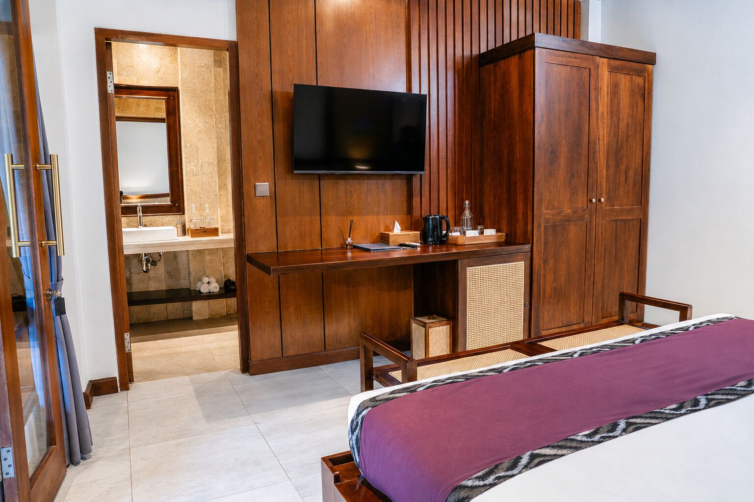 A hotel room with a bed, a TV mounted on the wall, a wooden wardrobe, and a small desk with a chair.