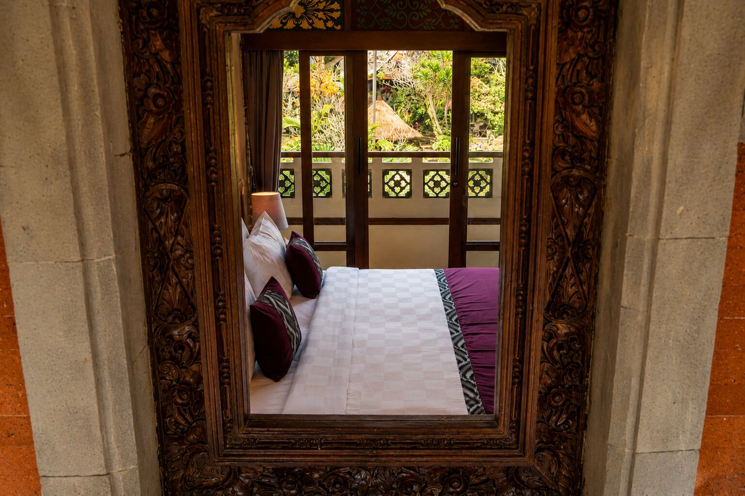 A bed with a checkered bedspread and pillows, framed by an ornate wooden structure with a view of a garden outside.