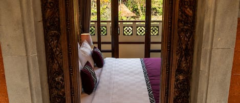 A bed with a checkered bedspread and pillows, framed by an ornate wooden structure with a view of a garden outside.