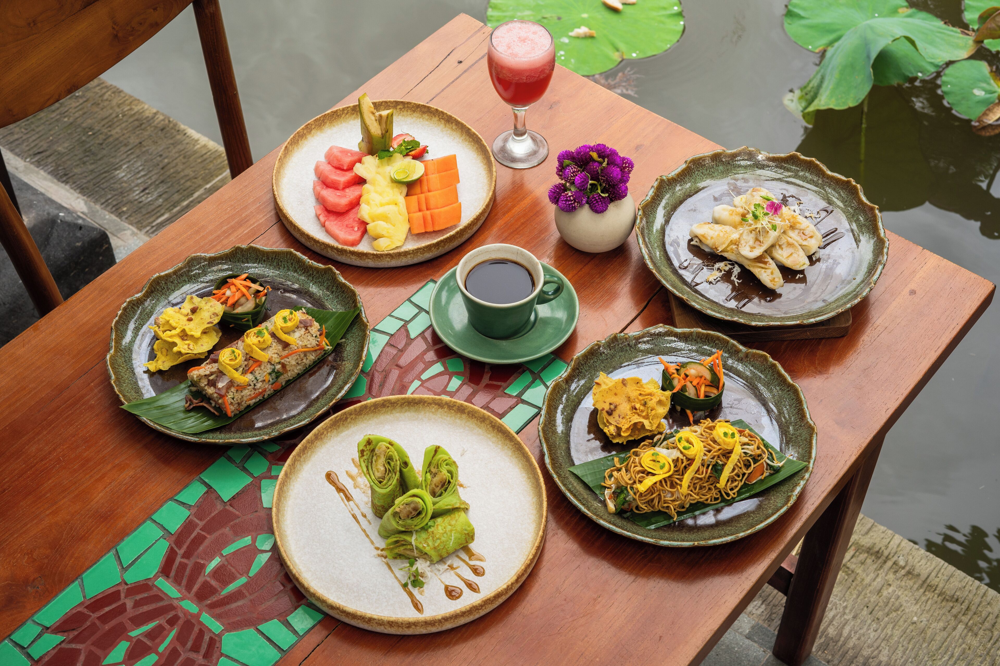A wooden table with various dishes, including a plate of sliced fruits, a bowl of noodles, and a cup of coffee.
