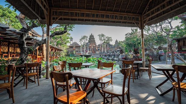 An outdoor seating area with wooden tables and chairs, overlooking a temple and a pond with lotus flowers. - Mumbul Ubud (Ubud)