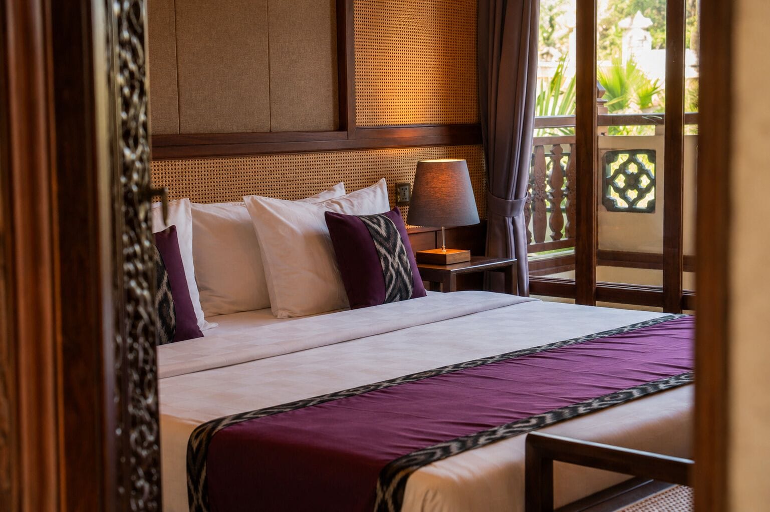 A hotel room with a bed, pillows, a nightstand, a lamp, and a view through a window.