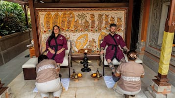 A group of people in traditional attire seated in a room with a large mural, a small table with bowls, and a statue.