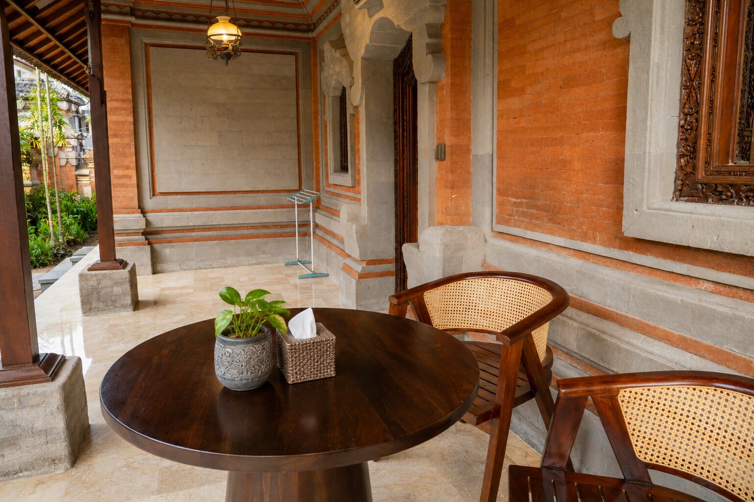 A round wooden table with a potted plant and a tissue box, surrounded by wicker chairs, in a room with brick walls and a tiled floor.