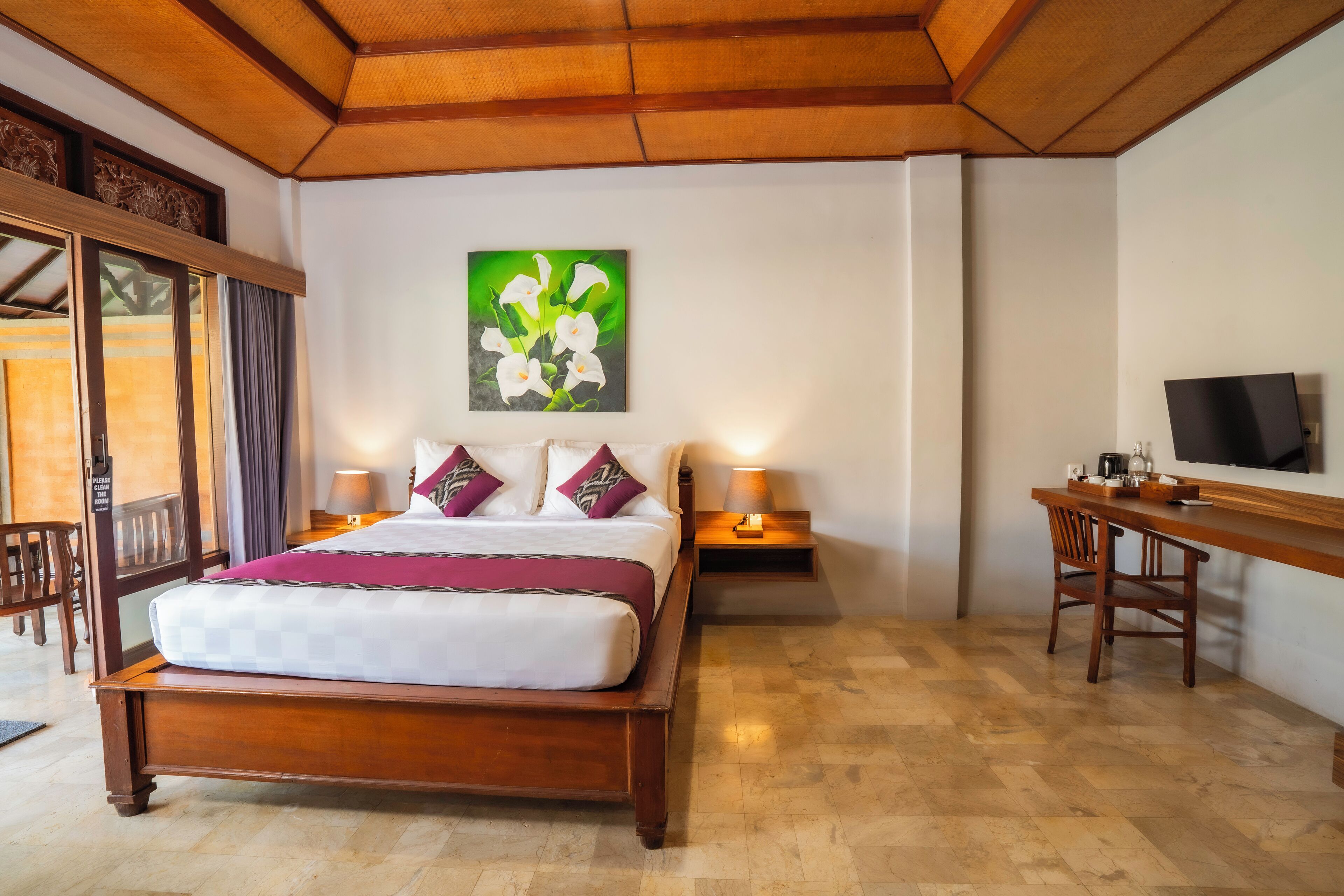 A hotel room with a wooden bed, a desk with a television, and a painting of flowers on the wall.