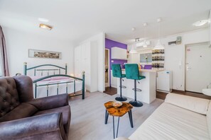 A modern room with a bed, a leather armchair, a small wooden table, and a kitchen area with a purple wall and white cabinets.