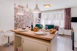A modern kitchen with a central island, pendant lights, a stone wall backsplash, and a dining area with a green chair and a television.