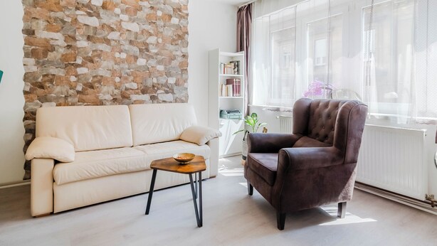 A living room with a stone wall, a white sofa, a brown armchair, a small wooden table, and a bookshelf. - Charming Downtown Studio, Zagreb, Croatia (Zagreb)