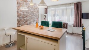 A modern kitchen with a central island, pendant lights, a stone accent wall, and a view of the living area with a TV and curtains. - Charming Downtown Studio, Zagreb, Croatia (Zagreb)