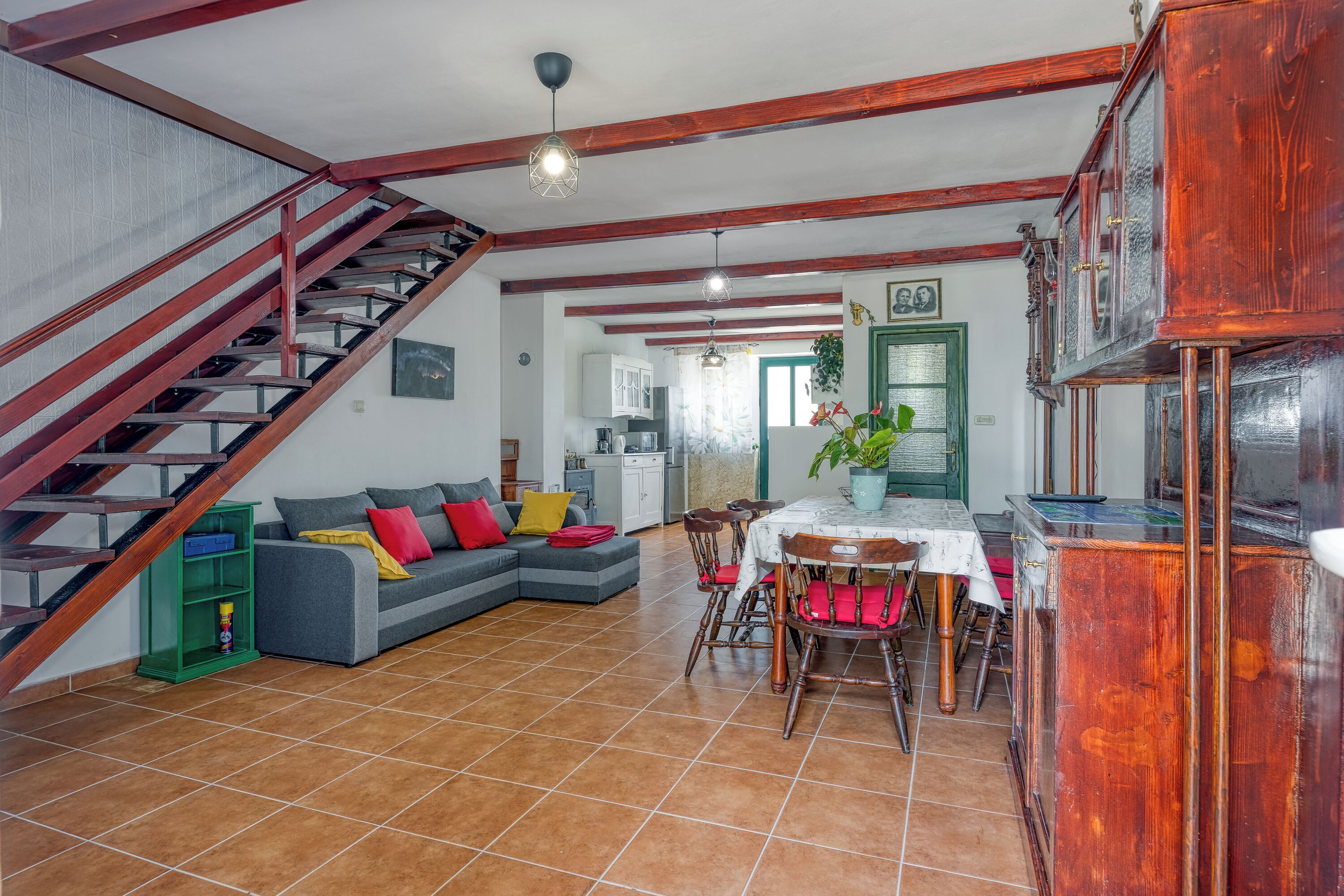 A spacious living area with a staircase, a dining table with chairs, a sofa with cushions, and a kitchen in the background.