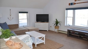 Un salon avec un canapé, une table basse blanche, un téléviseur posé sur un meuble blanc, une plante et une fenêtre donnant sur un beau paysage.