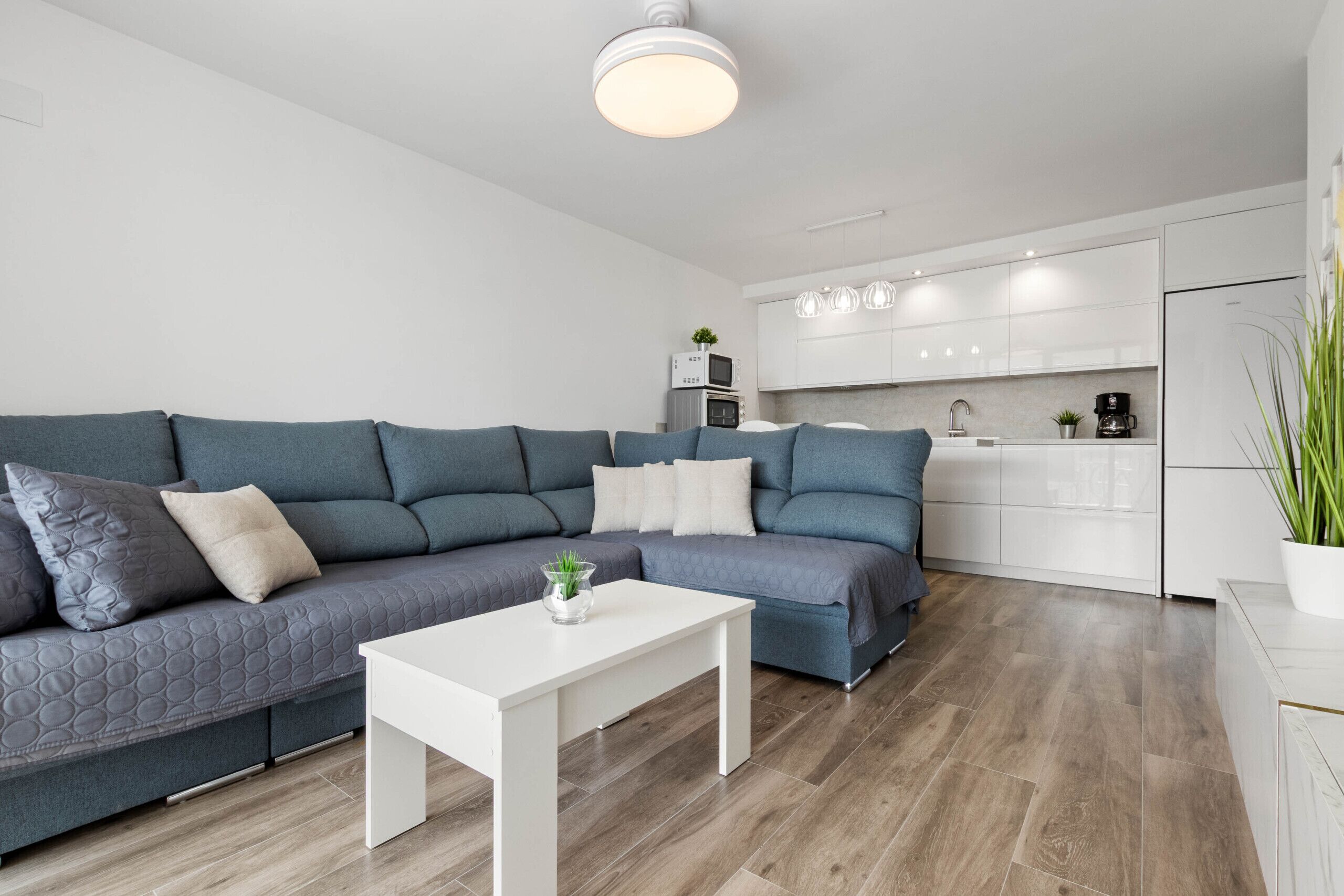 Un salón moderno con un sofá seccional gris, una mesa de centro blanca y una cocina al fondo.