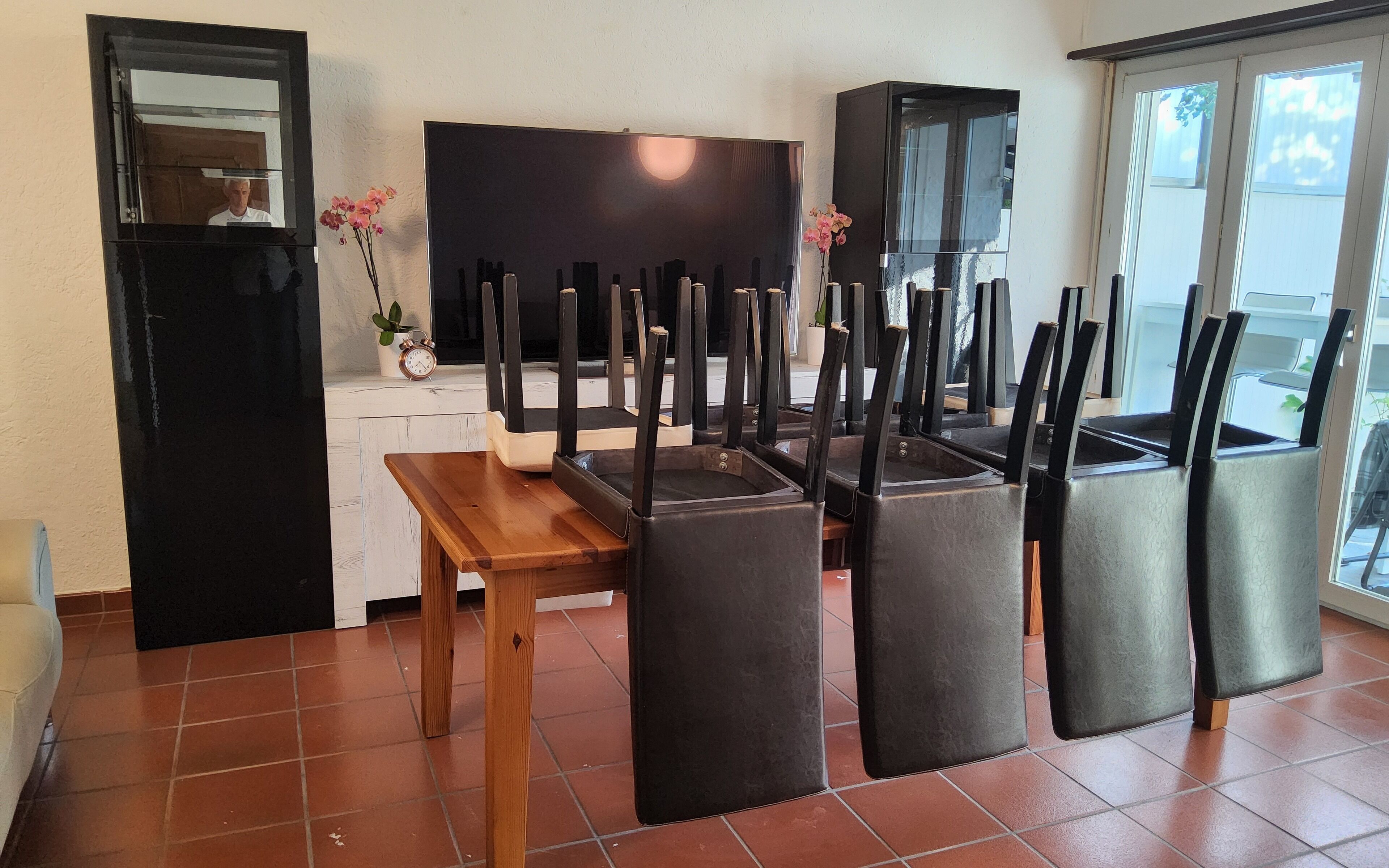 A dining area with a wooden table and black chairs, a TV, and a kitchen area with a refrigerator and cabinets.