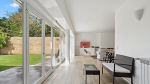 Un interior moderno con un amplio ventanal corredera que da a un jardín, un sofá, una mesa de comedor y un televisor al fondo.
