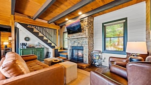 A cozy living room with a stone fireplace, leather sofas, a staircase, and a kitchen area.