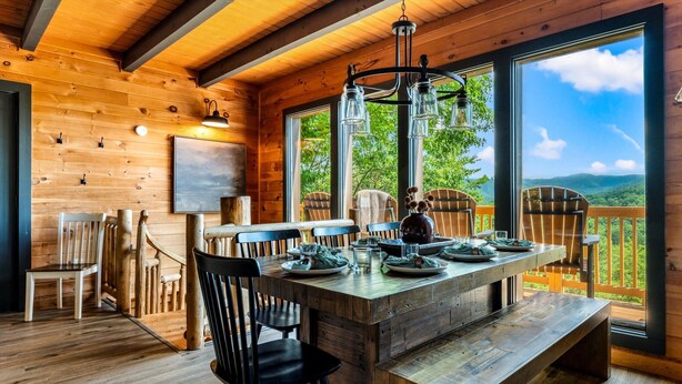 A wooden cabin interior with a dining table, chairs, and a large window offering a view of the outdoors.