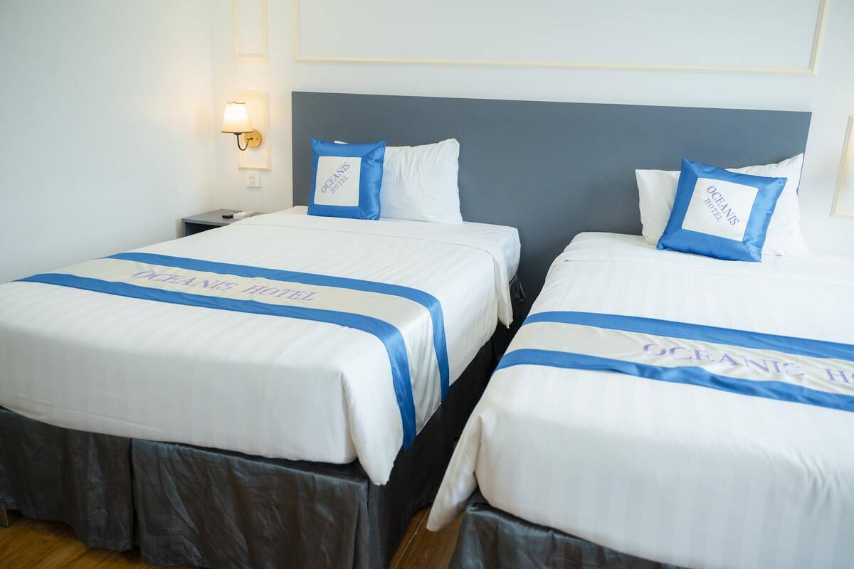 Two beds with matching blue and white bedding, a nightstand with a lamp, and a wall-mounted light fixture.