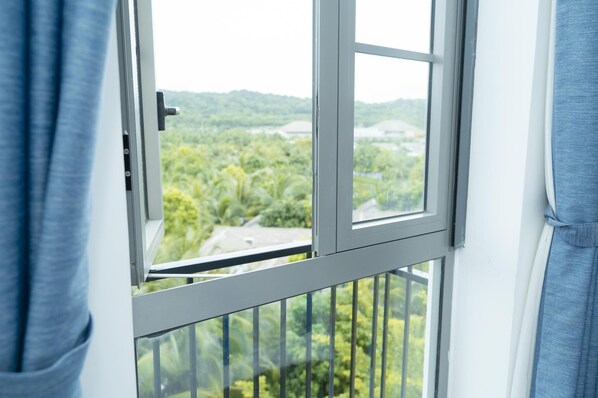 A window with a view of a green landscape, a metal railing, and a white wall. - Oceanis Hotel (Phu Quoc)