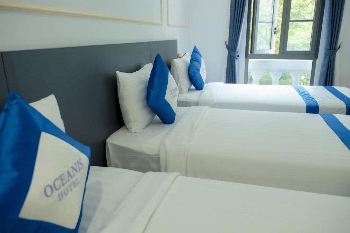 A hotel room with two beds, each with white bedding and blue pillows. The room has a grey headboard and a window with curtains.
