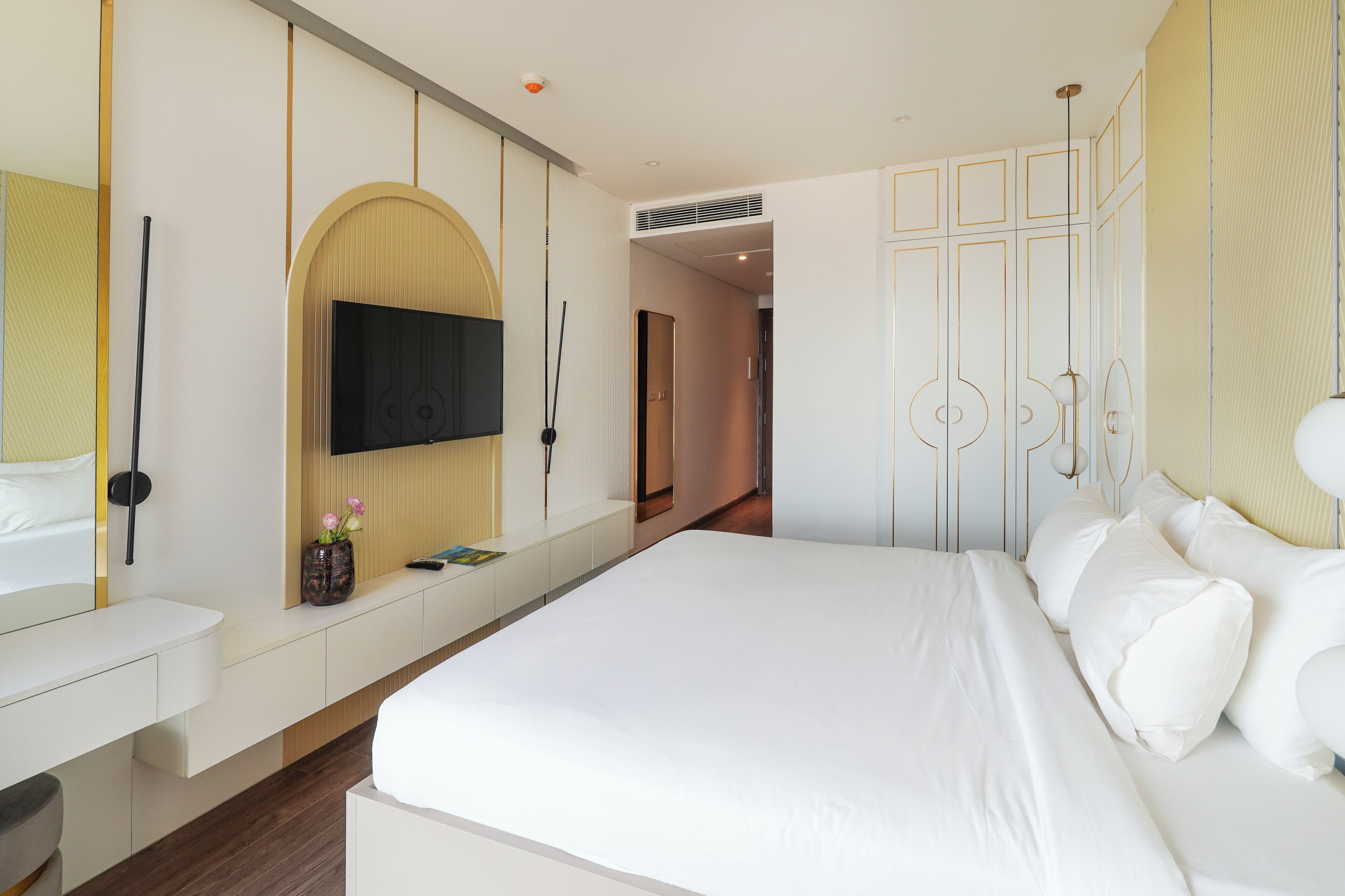 A modern hotel room with a large bed, a flat-screen TV mounted on the wall, a bedside table with a vase of flowers, and a wardrobe with a unique geometric design.