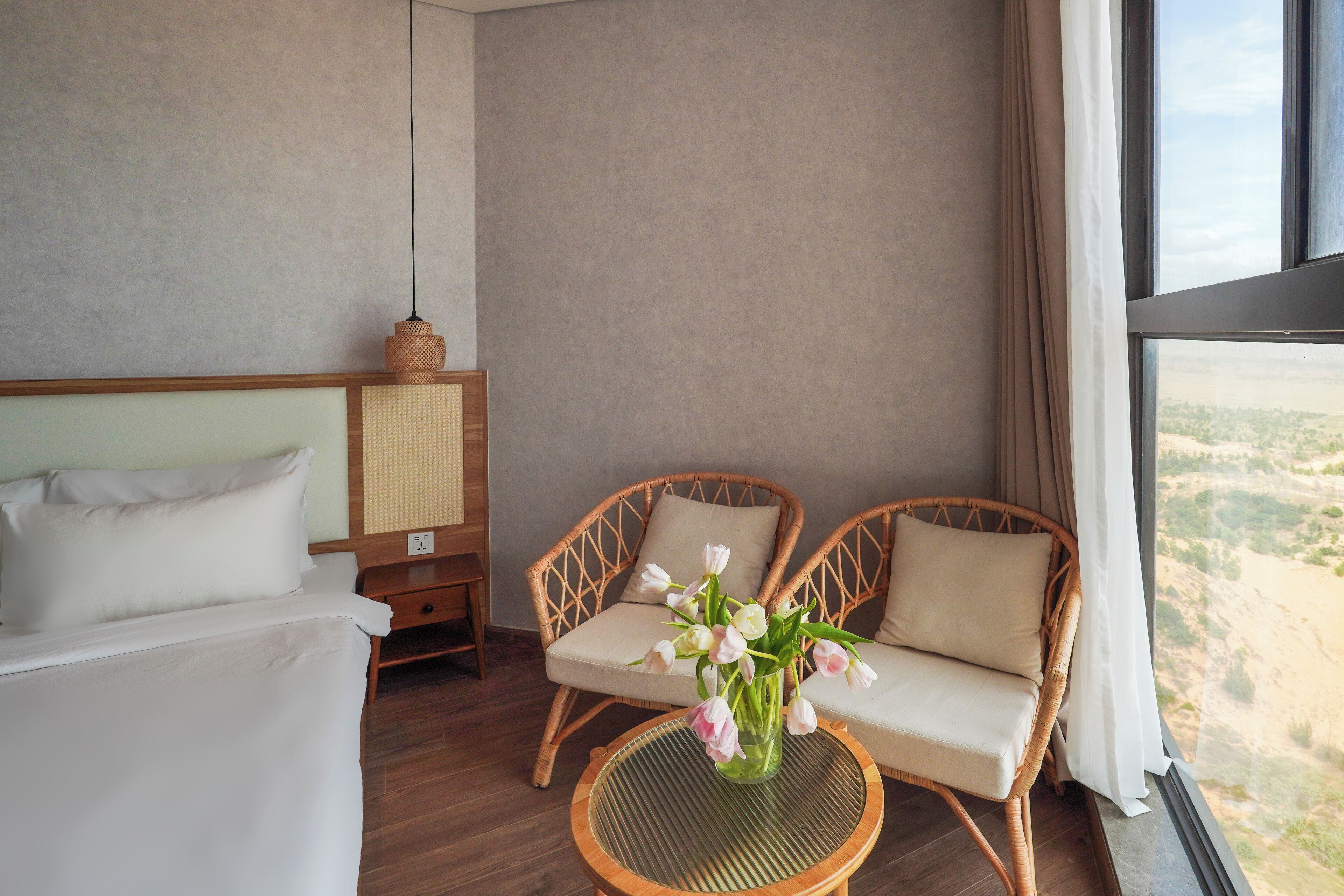 A hotel room with a bed, two wicker chairs, a small round table with a vase of flowers, and a large window with a view of the countryside.