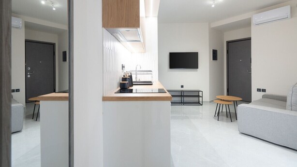 A modern, minimalist living space with a kitchen area, a flat-screen TV mounted on the wall, and a grey sofa. - LIV - Comfortable Athens Apartment S8-1B (Athens)
