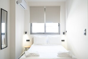 A hotel room with a bed, a window with blinds, a small table, and a wall-mounted air conditioner. - LIV - Comfortable Athens Studio S8-1A (Athens)