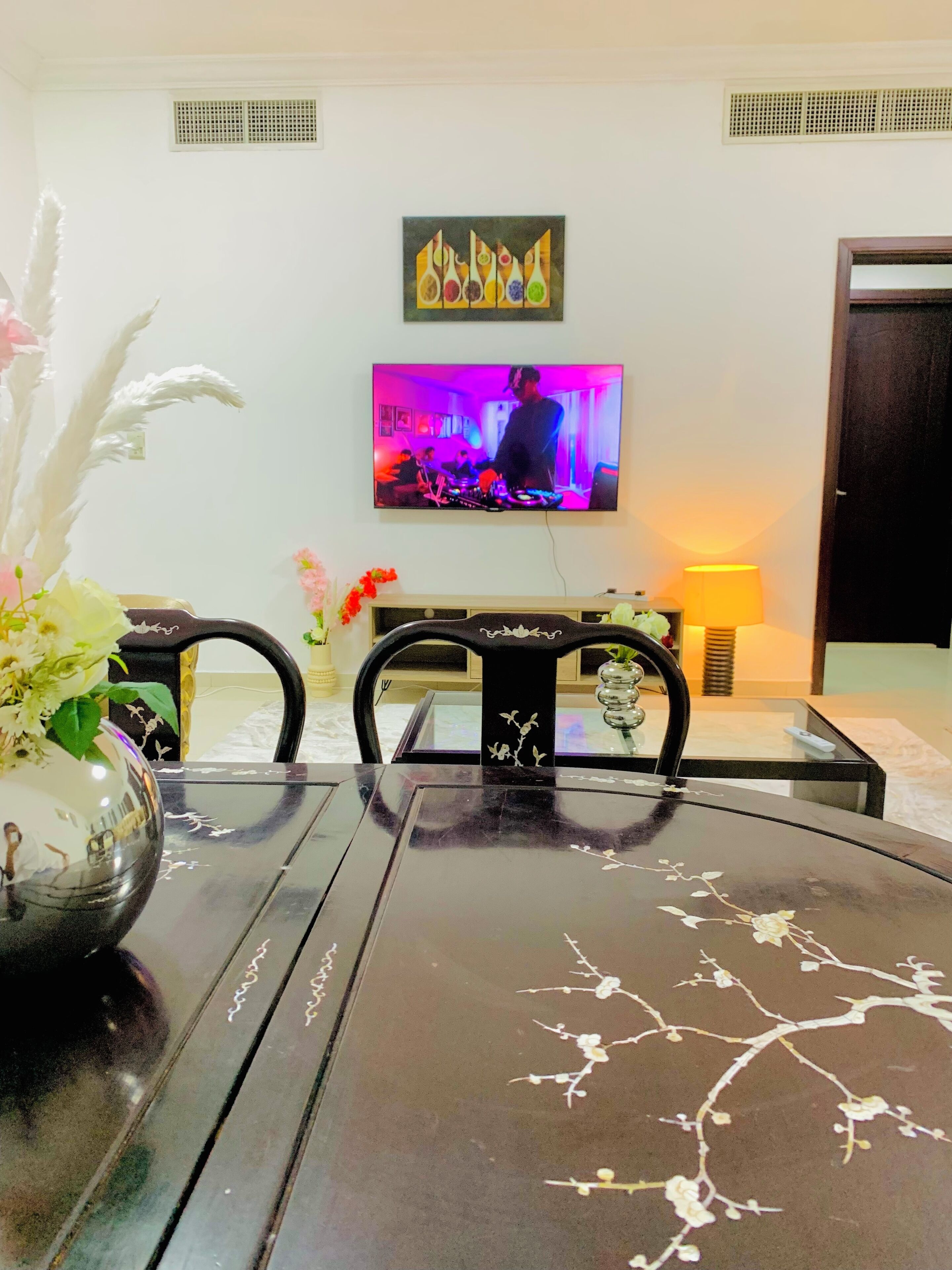 Un comedor con una mesa, sillas, un florero con flores, un televisor y una obra de arte colgada en la pared.