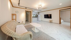 Een woonkamer met een rieten bank, een kleine witte tafel, een flatscreen televisie en een keuken op de achtergrond.