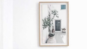 Une photographie en noir et blanc, encadrée, représentant une scène de rue avec un bâtiment et un arbre, un objet cylindrique vert et une lampe blanche posée sur un plancher en bois.