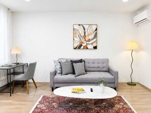 A modern living room with a grey sofa, a round coffee table, a patterned rug, a painting on the wall, and a floor lamp.