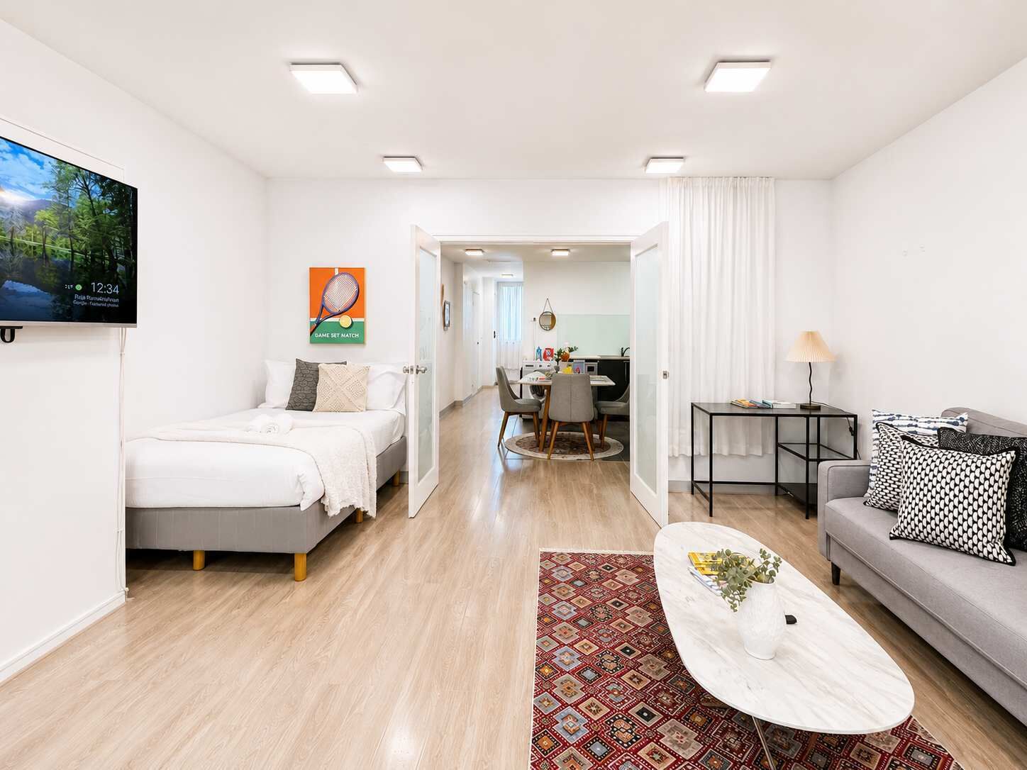 A modern hotel room with a bed, a sofa, a dining area, and a TV mounted on the wall.