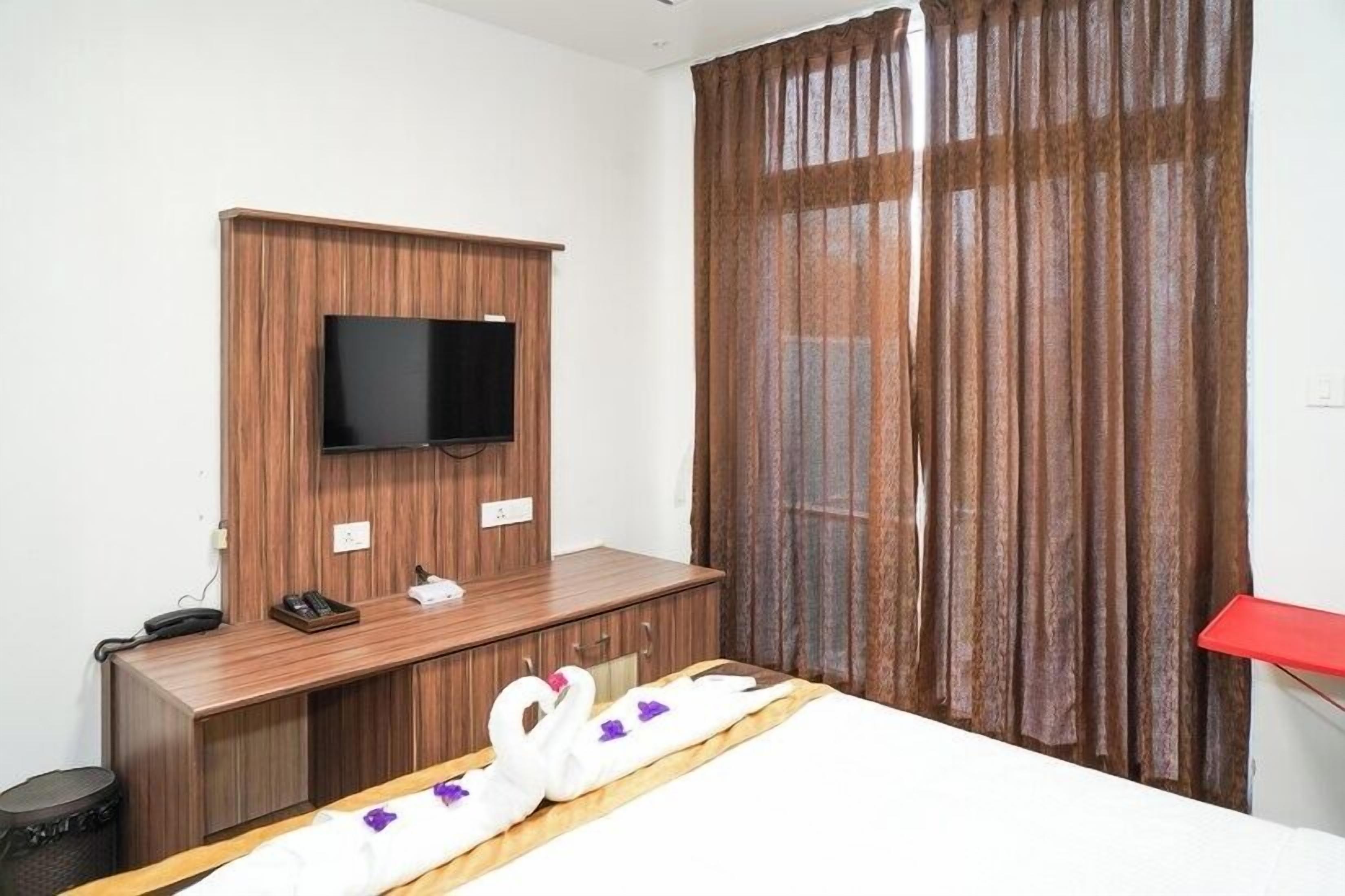 A hotel room with a wooden desk, a flat-screen TV mounted on the wall, a bed with a red bedside table, and a window with brown curtains.