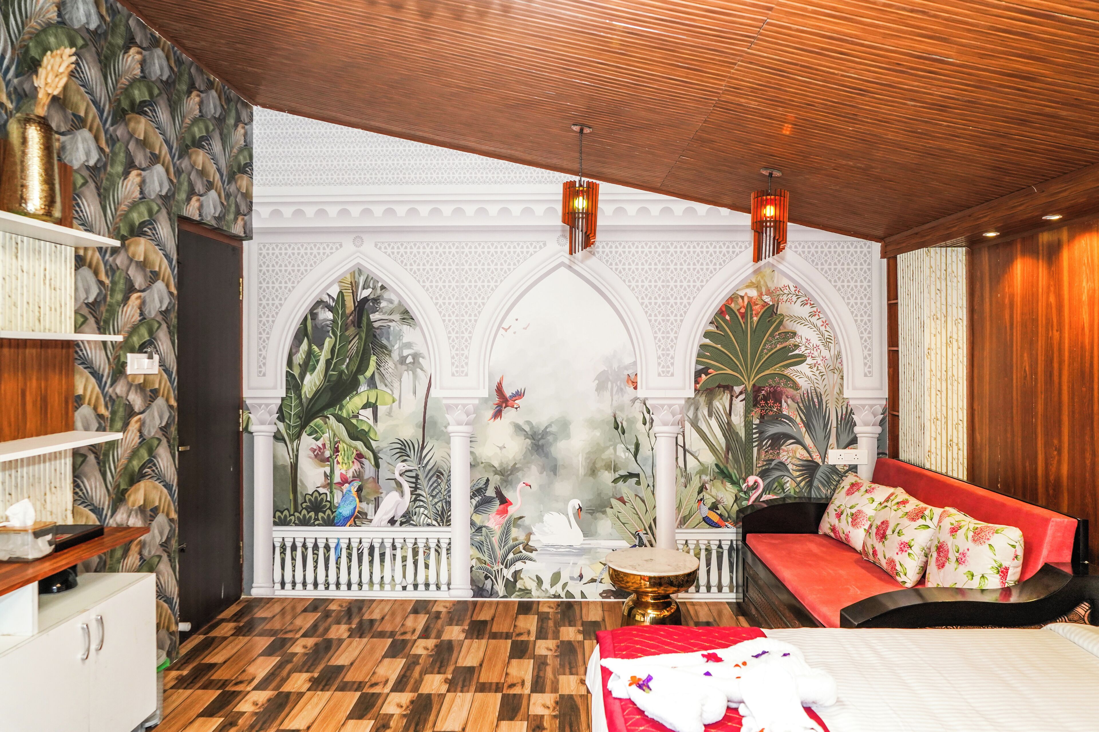 Una habitación con piso de madera a cuadros, un sofá rojo con cojines florales, una cama blanca y una pared decorativa con murales tropicales.