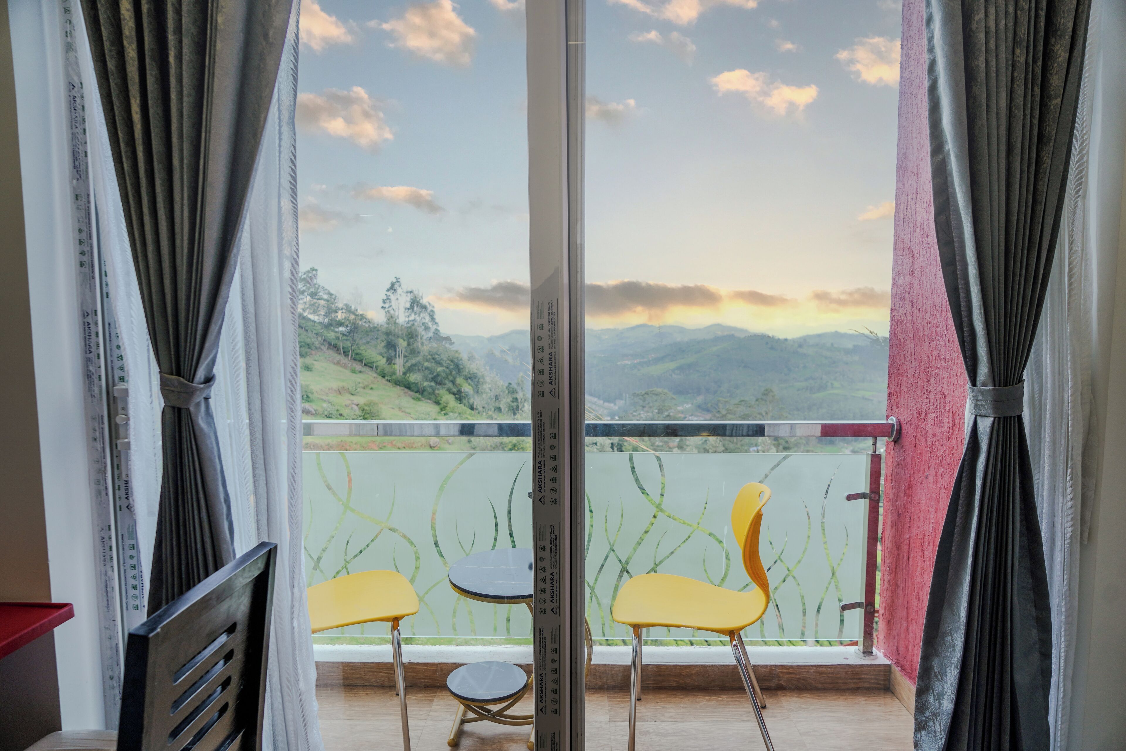 Un balcone con due sedie gialle e un tavolino, affacciato su un paesaggio pittoresco con colline e cielo sereno.