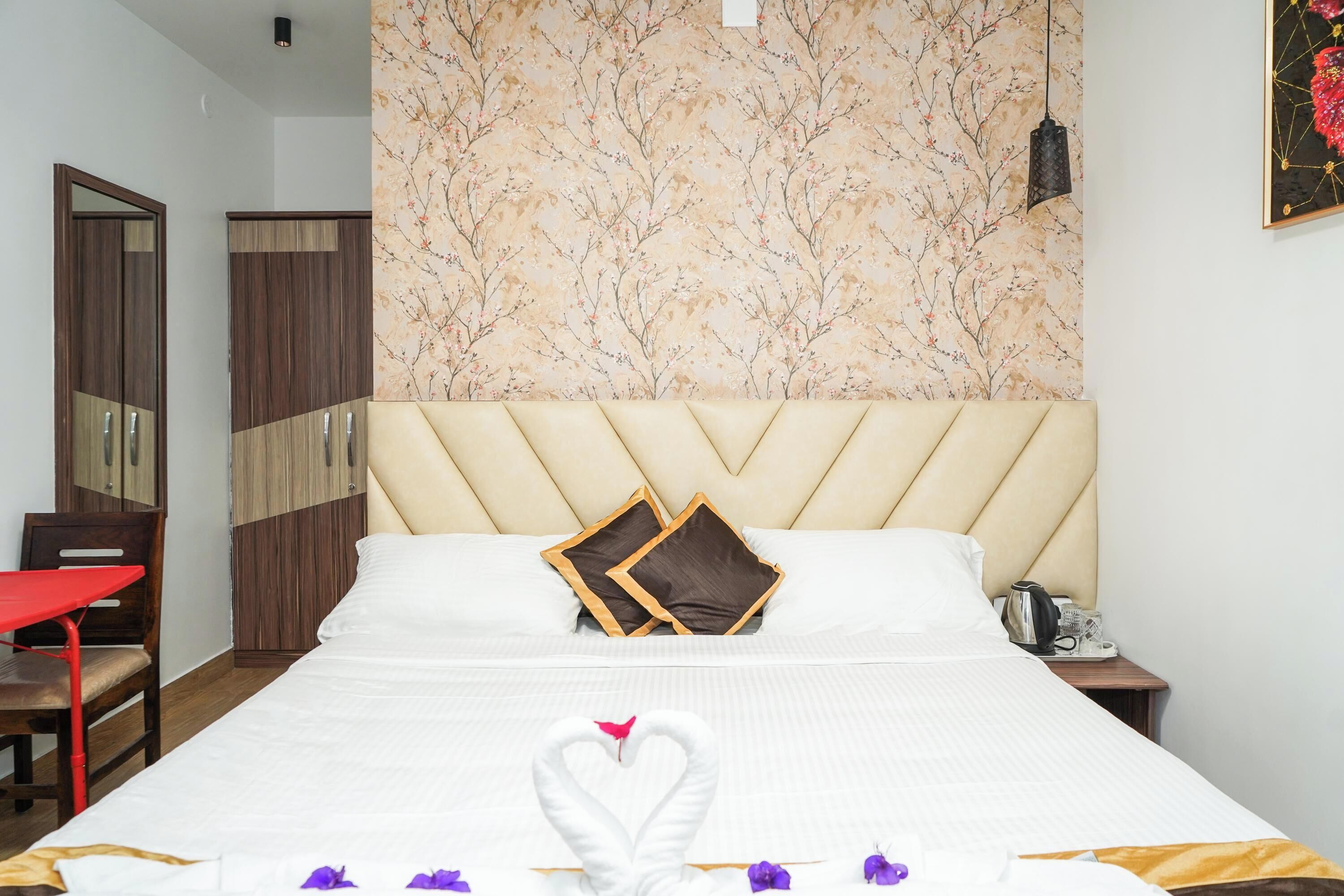 Habitación de hotel con una cama grande, cabecera, mesita de noche, una silla roja, armario y papel tapiz floral.