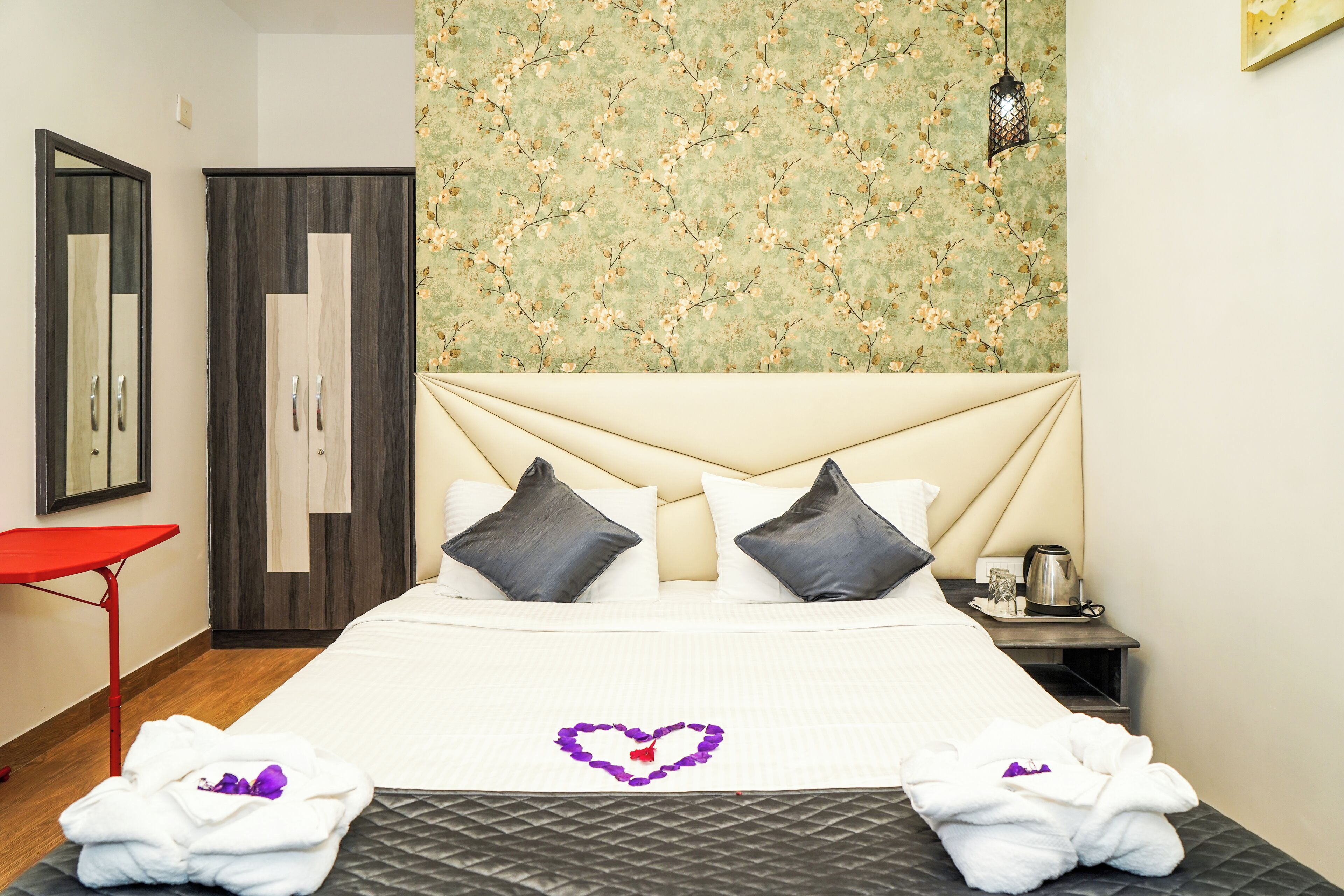 A hotel room with a bed, two pillows, a nightstand, a red chair, and a wall with a floral pattern.