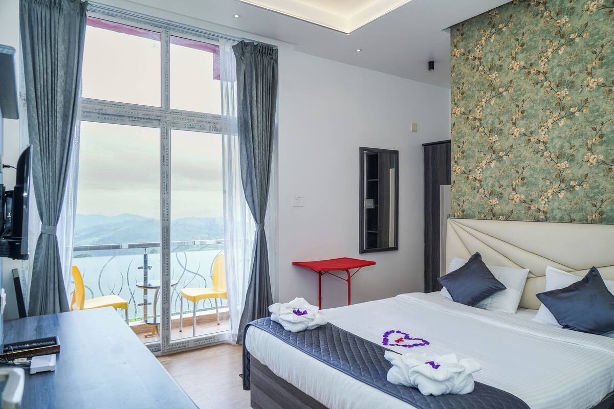 Une chambre d’hôtel avec un grand lit, une table de chevet rouge, un balcon avec des chaises jaunes et une vue panoramique.