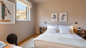 A bedroom with a large bed, two bedside tables, a desk with a chair, and a window with a view of buildings.