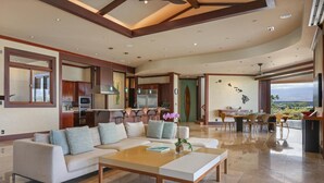 Amplia sala de estar con un sofá grande, una mesa de centro y un comedor con sillas. Se observa una cocina con electrodomésticos modernos y vistas al exterior.