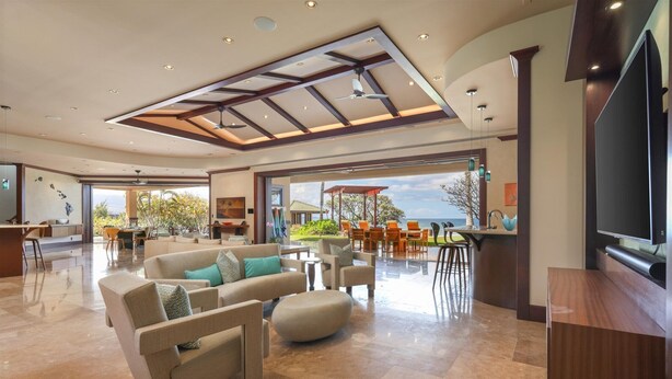 Amplia sala de estar con un sofá grande, una mesa de centro de madera y un televisor sobre un mueble de madera. Hay un comedor con sillas color naranja y vistas al exterior.