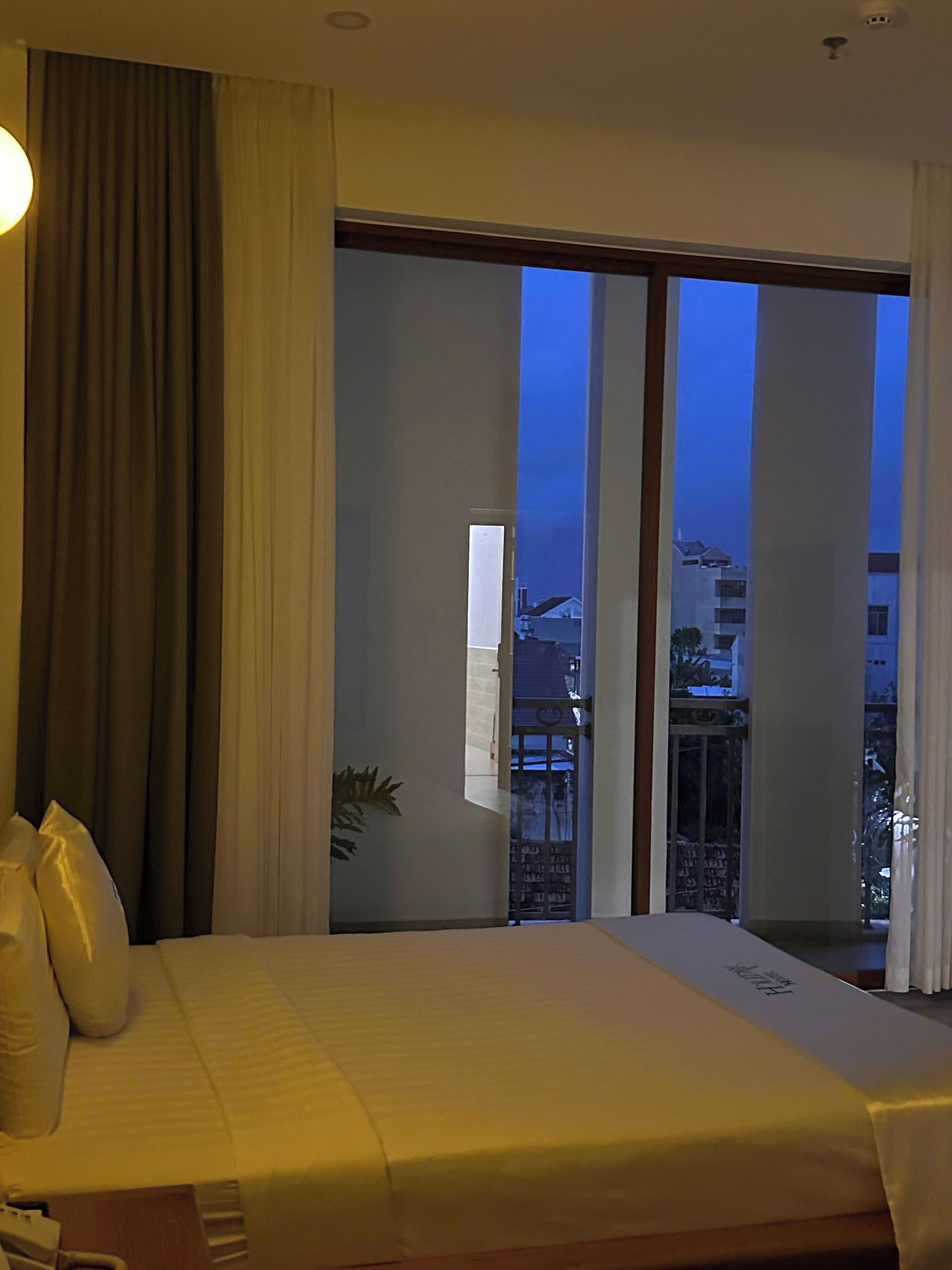 A hotel room with a bed, curtains, a window with a view of buildings, and a plant.
