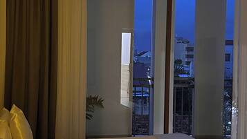 A hotel room with a bed, curtains, a window with a view of buildings, and a plant.
