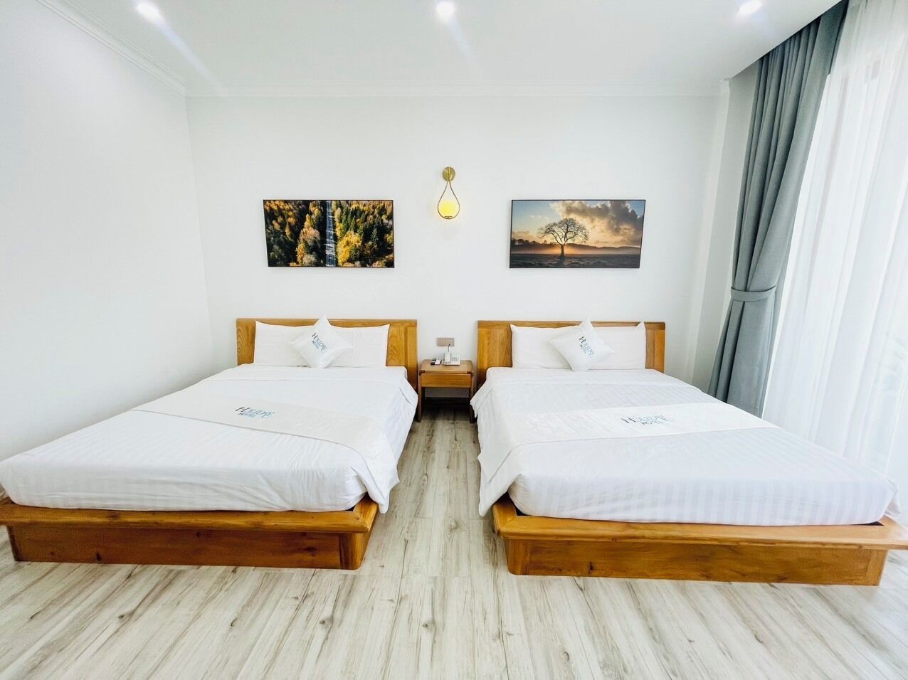 Una habitación de hotel con dos camas de madera, ropa de cama blanca, una mesita de noche, una lámpara y dos cuadros en la pared.